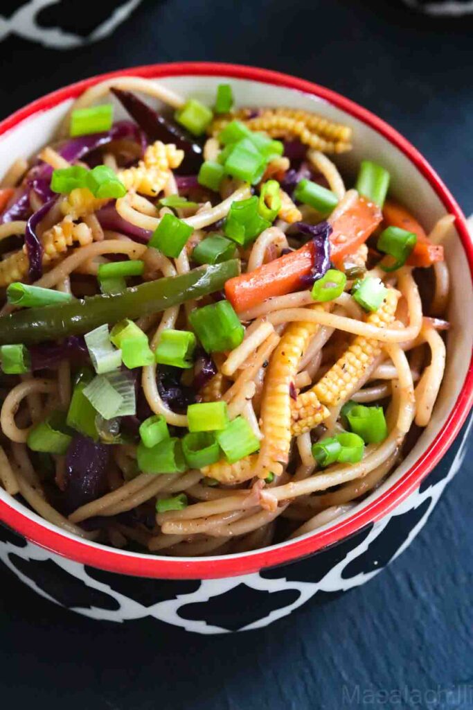 noodles with mixed vegetables garnished with spring onions served in a white and black bowl with a red rim