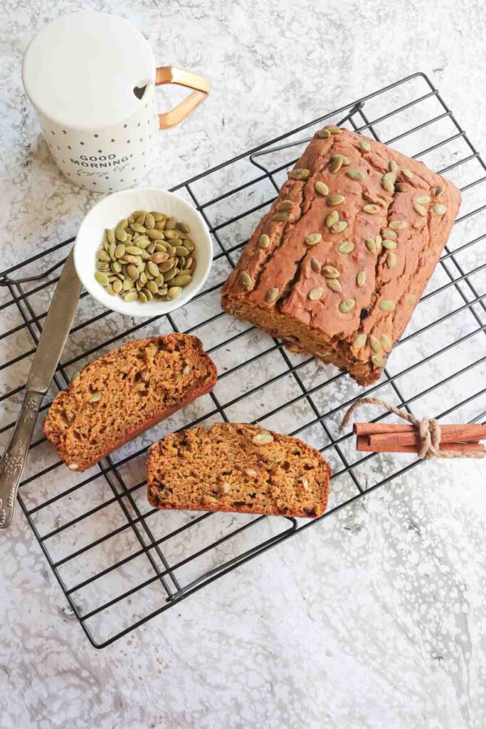 2 slices of pumpkin bread with a half cut loaf of pumpkin bread placed on a wire rack with cinnamon stick and pepitas in a small white bowl