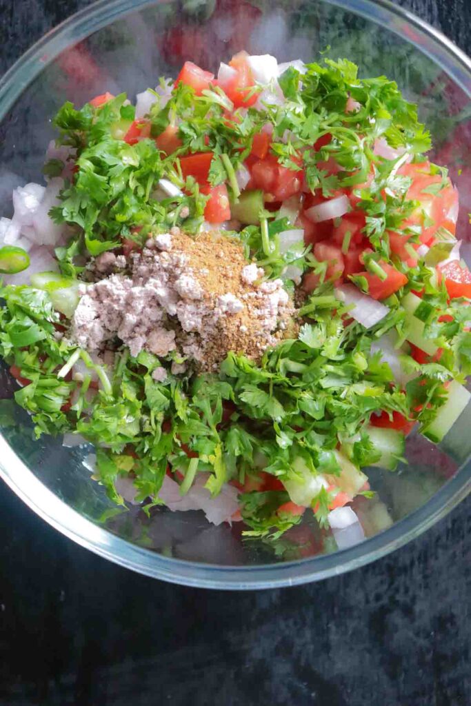 Ingredients for kachumber salad added to a glass bowl with coriander leaves, rock salt and roasted cumin powder